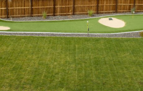 A backyard landscape featuring a well-manicured putting green with sand trap elements, bordered by gravel and a wooden fence. The green area is complemented by a lush grass lawn and scattered shrubs, providing a functional and aesthetically pleasing outdoor sports area.